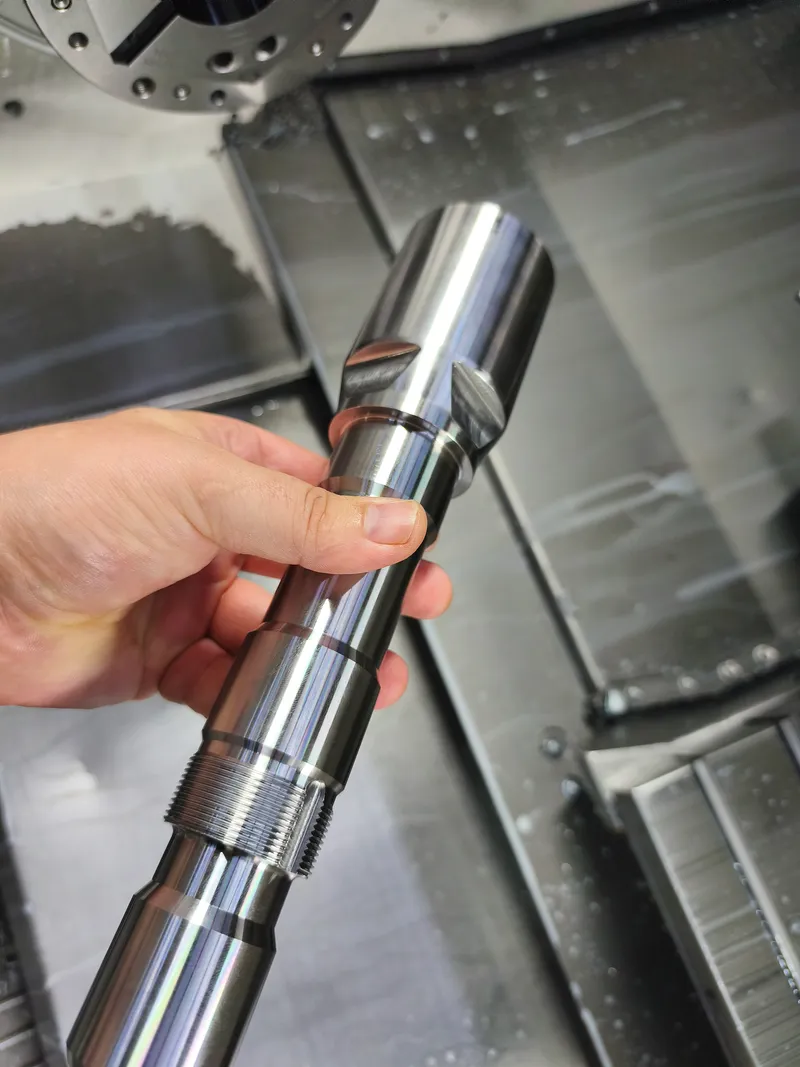Gedrehte Welle mit Absätzen und Gewinde, in der Hand vor der Drehmaschine